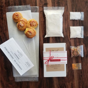 Op de afbeelding: Overzicht van een Zweedse Limpa brood bakset. De set bevat voorgebakken broodjes, bloem, suiker, gist, rozijnen, kruiden en instructies. De ingrediënten zijn gerangschikt op een houten oppervlak.