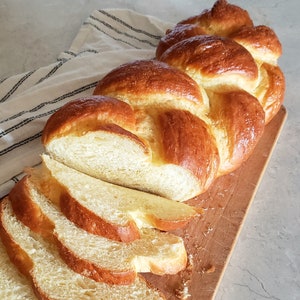 Puede incluir: Una hogaza de pan trenzado cortada en una tabla de cortar de madera. El pan es de color marr&oacute;n dorado y tiene una textura suave y esponjosa.