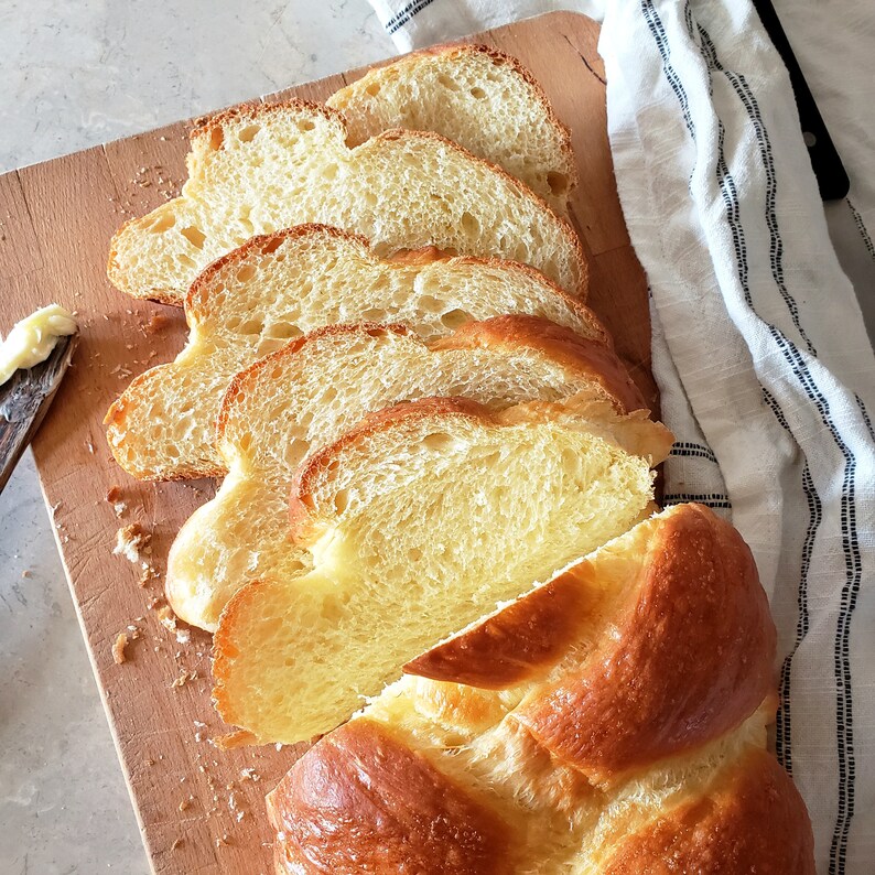 Puede incluir: Una hogaza de pan reci&eacute;n horneado cortada, revelando un interior suave y dorado. El pan est&aacute; dispuesto sobre una tabla de cortar de madera con una servilleta de tela blanca en el fondo.