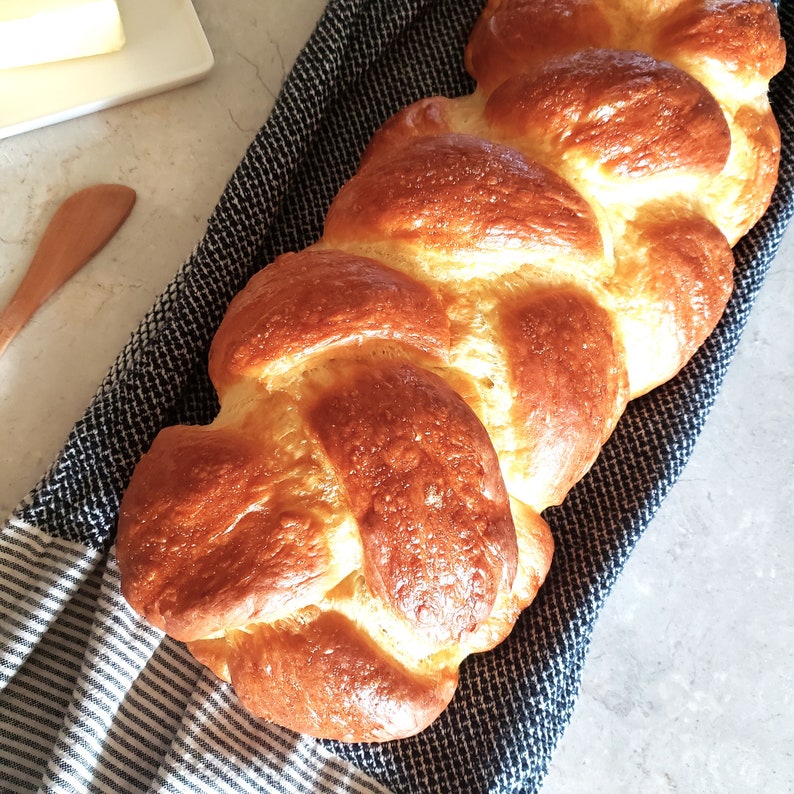 Puede incluir: Una hogaza de pan trenzado con una corteza dorada, sentada sobre una toalla de cocina a rayas azules y blancas.