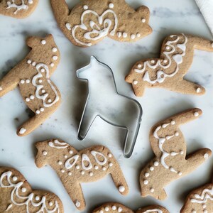 May include: A close-up of a metal cookie cutter shaped like a Dala horse, surrounded by gingerbread cookies decorated with white icing. The cookies are on a white marble surface.