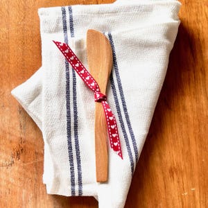 Pu&ograve; includere: Un coltello da burro in legno con un nastro rosso legato intorno. Il nastro ha cuori bianchi stampati. Il coltello &egrave; su un asciugamano da cucina bianco con strisce blu.