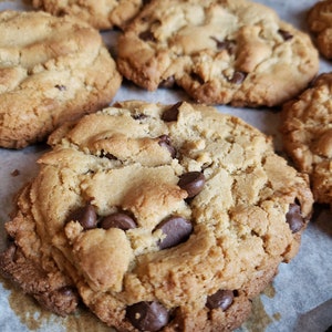 Pu&ograve; includere: Primo piano di una teglia di biscotti al cioccolato appena sfornati su una teglia da forno rivestita di carta da forno. I biscotti sono dorati e hanno un esterno leggermente croccante con un interno morbido e gommoso. Le gocce di cioccolato sono sciolte e leggermente distribuite, aggiungendo un sapore ricco e decadente ai biscotti.