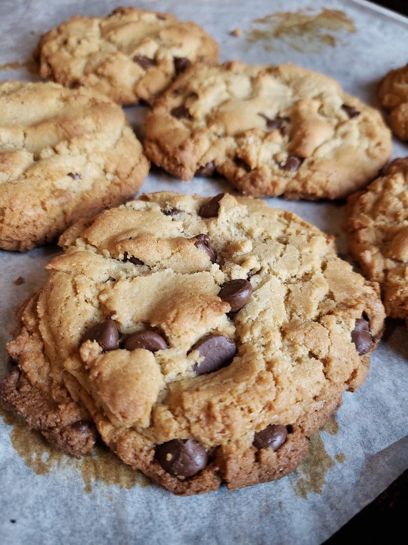 Pu&ograve; includere: Primo piano di una teglia di biscotti al cioccolato appena sfornati su una teglia da forno rivestita di carta da forno. I biscotti sono dorati e hanno un esterno leggermente croccante con un interno morbido e gommoso. Le gocce di cioccolato sono sciolte e leggermente distribuite, aggiungendo un sapore ricco e decadente ai biscotti.