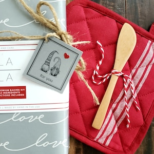Pu&ograve; includere: Un'etichetta regalo grigia con due gnomi che si tengono per mano e un cuore rosso, legata con uno spago a una scatola regalo grigia e bianca. Il testo sull'etichetta dice "per te".