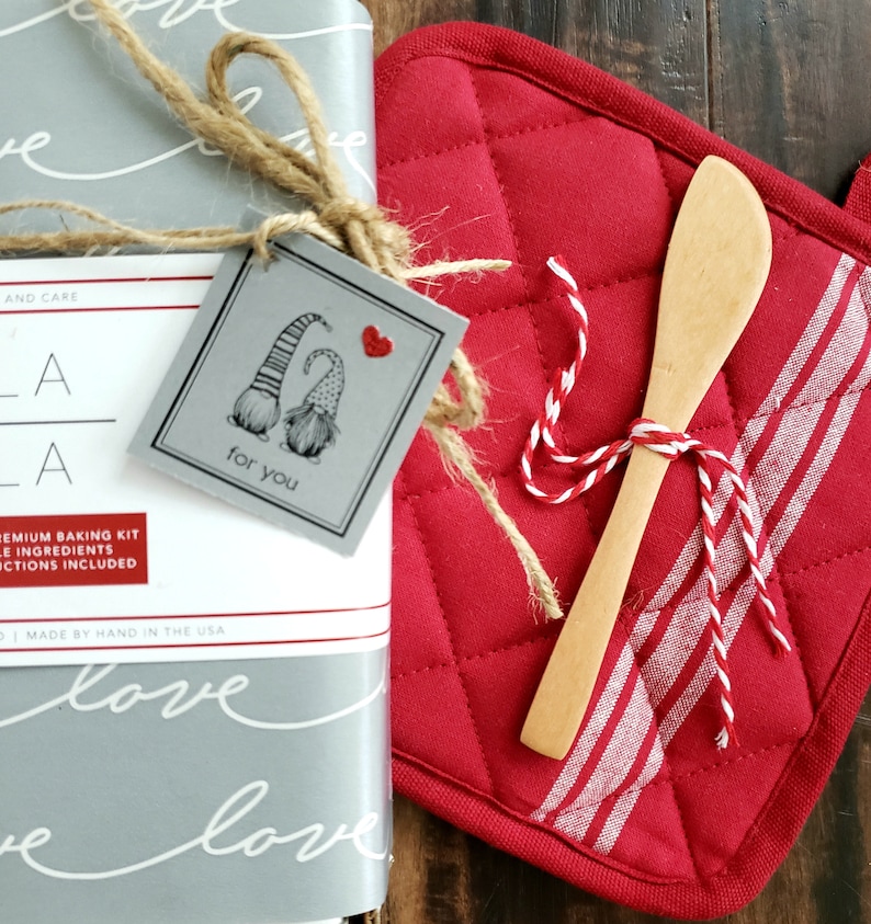 Pu&ograve; includere: Un'etichetta regalo grigia con due gnomi che si tengono per mano e un cuore rosso, legata con uno spago a una scatola regalo grigia e bianca. Il testo sull'etichetta dice "per te".