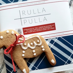 May include: A gingerbread horse cookie with white icing and a red twine bow rests on a baking kit box. The box has a blue and white plaid pattern and the text "RULLA PULLA" and "HANDMADE BY YOU | PREMIUM BAKING KIT".