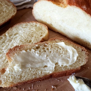 Puede incluir: Primer plano de una rebanada de pan con mantequilla untada encima. El pan es de color marr&oacute;n claro y tiene una textura suave y esponjosa. La mantequilla es blanca y cremosa.
