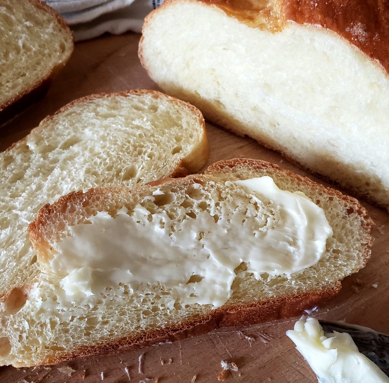 Puede incluir: Primer plano de una rebanada de pan con mantequilla untada encima. El pan es de color marr&oacute;n claro y tiene una textura suave y esponjosa. La mantequilla es blanca y cremosa.