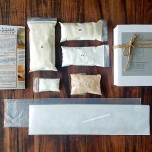 Op de afbeelding: Een bakpakket met diverse ingrediënten, waaronder zakken bloem met de labels A, B, C en D, gist en een doos met kardemom broodjes. De set is vastgebonden met touw en bevat instructies.