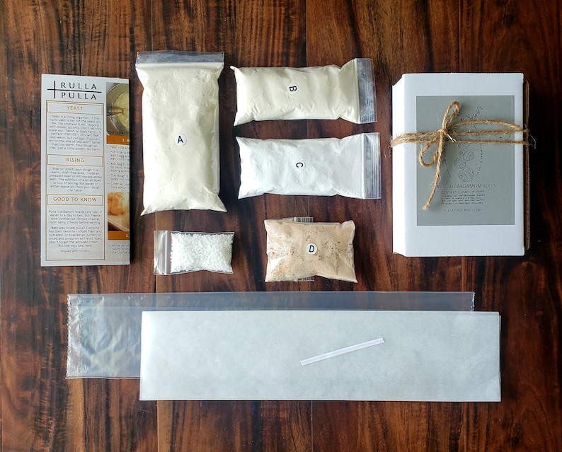 May include: A baking kit with various ingredients, including bags of flour labeled A, B, C, and D, yeast, and a box of cardamom rolls. The kit is tied with twine and includes instructions. The items are arranged on a wooden surface.