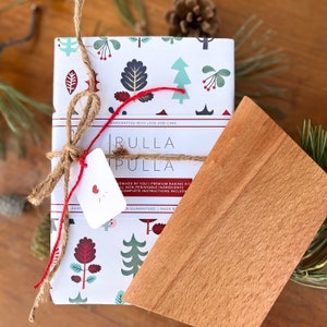 May include: A wrapped baking kit with a wooden scraper. The kit is wrapped in white paper with a repeating pattern of trees and leaves in teal, burgundy, and gray. It is tied with twine and red string, and includes a tag with a heart.