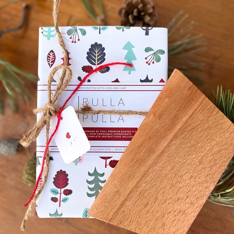 May include: A wrapped baking kit with a wooden scraper. The kit is wrapped in white paper with a repeating pattern of trees and leaves in teal, burgundy, and gray. It is tied with twine and red string, and includes a tag with a heart.