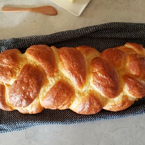 Puede incluir: Una hogaza de pan trenzado, de color marr&oacute;n dorado y brillante con un brillo de aceite, se encuentra sobre una toalla de cocina a rayas azules y blancas.
