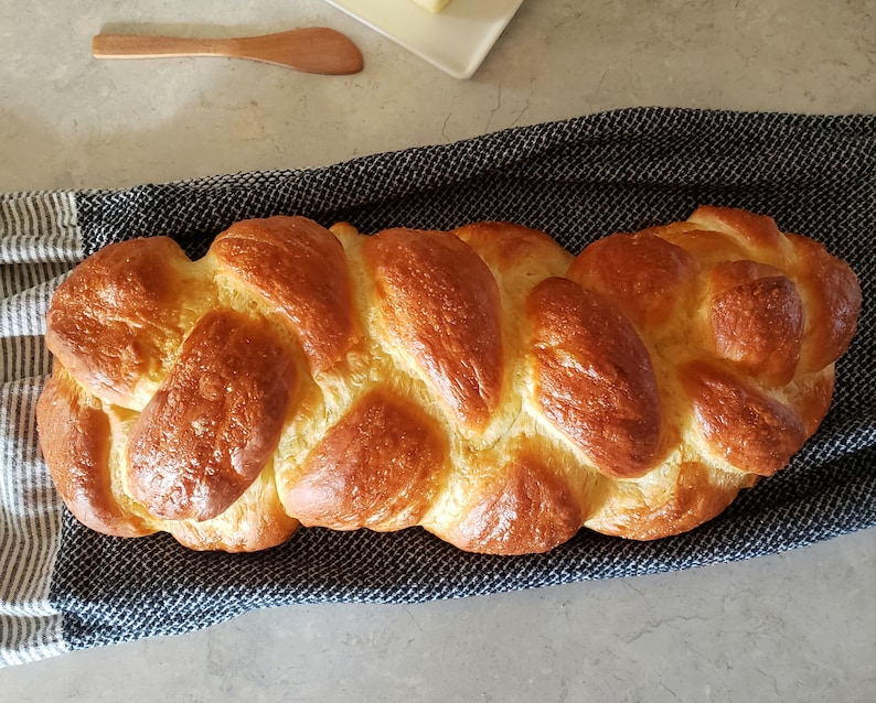 Puede incluir: Una hogaza de pan trenzado, de color marr&oacute;n dorado y brillante con un brillo de aceite, se encuentra sobre una toalla de cocina a rayas azules y blancas.