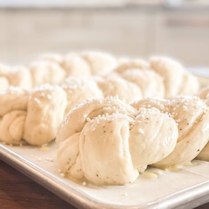 May include: Close-up of several unbaked, braided loaves of bread on a metal baking sheet. The dough is a light beige color, with visible flecks of herbs and coarse salt sprinkled on top. The sheet sits on a wooden surface.