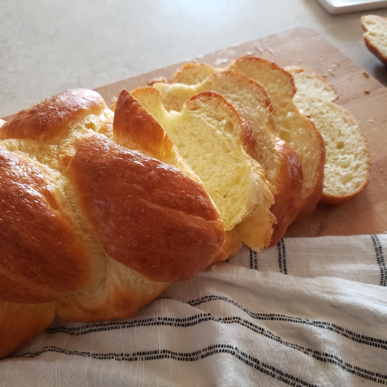 Puede incluir: Una hogaza de pan trenzado cortada en una tabla de cortar de madera. El pan es de color marr&oacute;n dorado y tiene una textura suave y esponjosa.