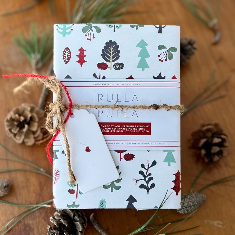 Pu&ograve; includere: Una scatola bianca con un design di albero di Natale rosso e verde. La scatola &egrave; avvolta con spago e un nastro rosso. Il testo sulla scatola recita "RULLA PULLA" e "HANDCRAFTED WITH LOVE AND CARE".