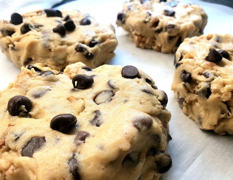 Pu&ograve; includere: Quattro palline rotonde di impasto per biscotti con gocce di cioccolato su una superficie di carta da forno bianca. L'impasto &egrave; di colore marrone chiaro e le gocce di cioccolato sono di colore marrone scuro.