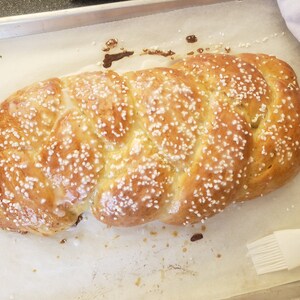 May include: A golden-brown, braided loaf of bread, sprinkled with white sugar crystals, sits on parchment paper in a baking pan. The bread has a glossy glaze and is ready to be served.
