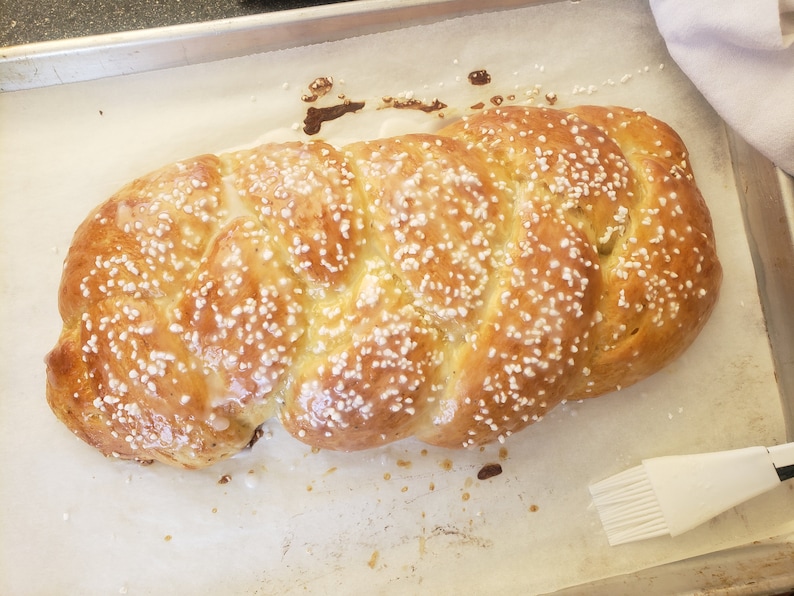 May include: A golden-brown, braided loaf of bread, sprinkled with white sugar crystals, sits on parchment paper in a baking pan. The bread has a glossy glaze and is ready to be served.