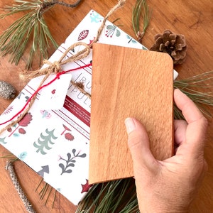 May include: A light-colored wooden cutting board with rounded corners is held over a package wrapped in decorative paper. The package is tied with twine and red string. Pine needles and a pine cone are scattered around the items.