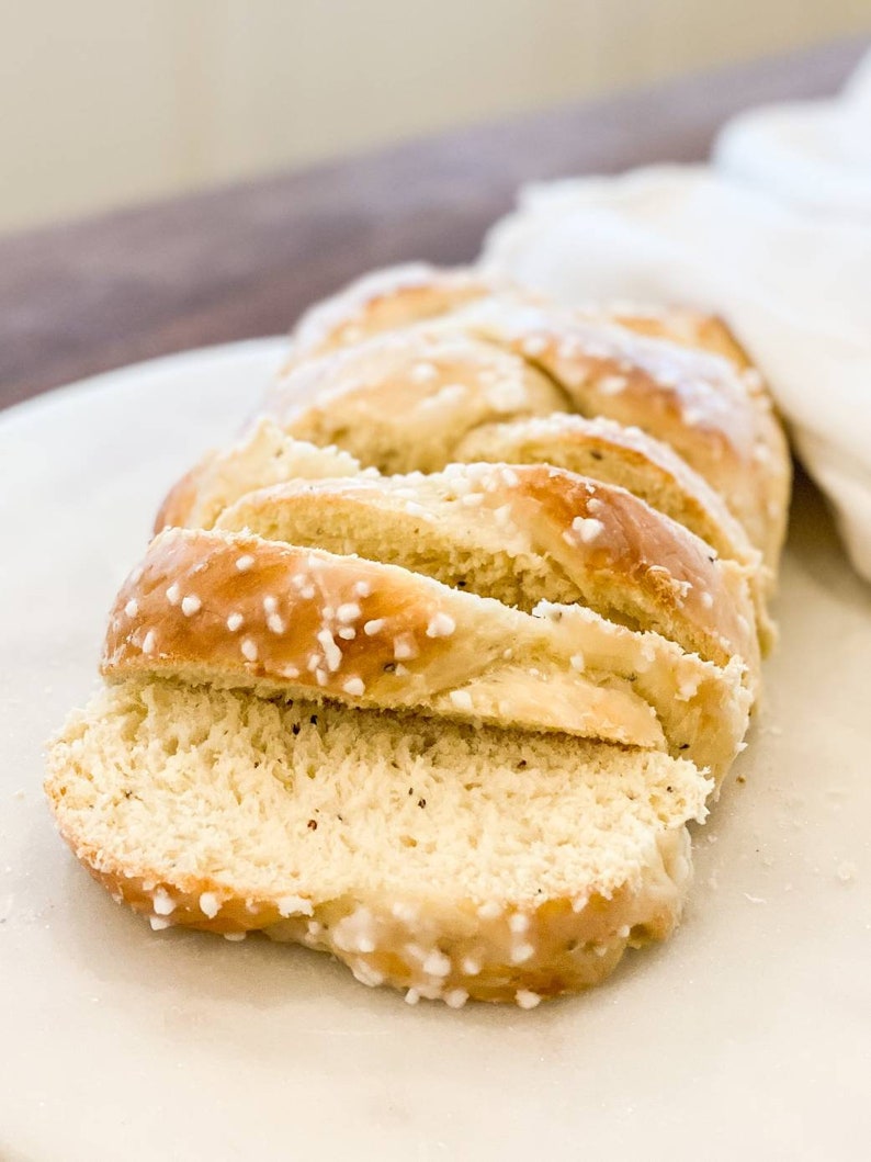 May include: Sliced loaf of challah bread with a golden crust and white, sugar-like sprinkles. The bread is on a white plate with a white cloth in the background. The bread is sliced and ready to eat.