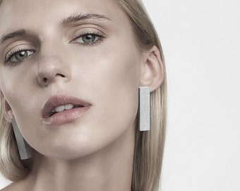 May include: A woman with blonde hair wearing a white sleeveless top and silver bar earrings.