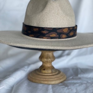 May include: A beige fedora hat featuring a wide, flat brim and a pinched crown. The hat is accented with a black fabric band decorated with a brown and black leopard print pattern that encircles the base of the crown above the brim.