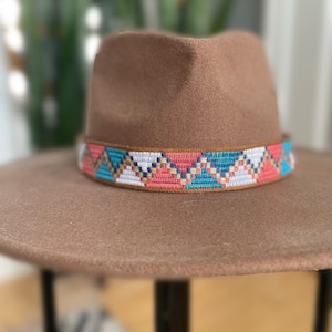 May include: A brown fedora hat featuring a wide, flat brim and a structured crown. The base of the crown is encircled by a decorative woven band displaying a repeating geometric zigzag pattern in shades of coral, teal, white, and gold.