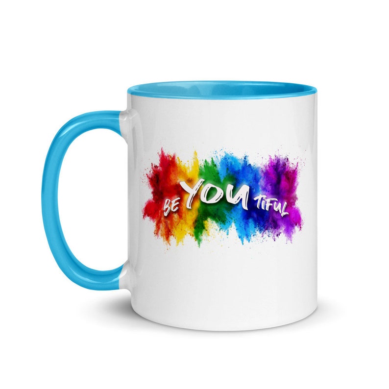 May include: White ceramic coffee mug with a light blue rim and handle. The mug features a rainbow-colored powder explosion with the text "BE YOU TIFUL" in white.