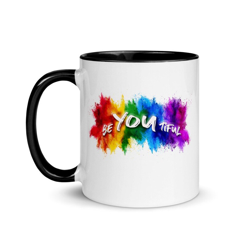 May include: White ceramic coffee mug with a black rim and handle. The mug features a rainbow-colored paint splatter design with the text "BE YOU TIFUL" in white.