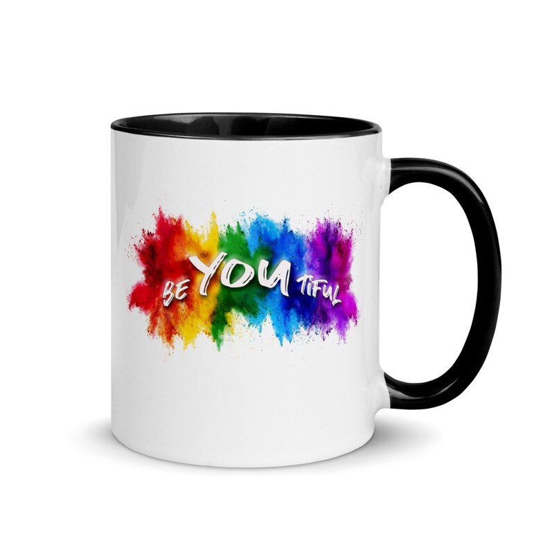 May include: White ceramic coffee mug with a black rim and handle. The mug features a colorful rainbow explosion with the text "Be You Tiful" in white.