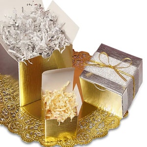 Pode incluir: Três caixas de presente douradas com detalhes prateados. As caixas estão abertas e cheias de papel de seda branco e creme. As caixas estão sobre um guardanapo de renda dourado.