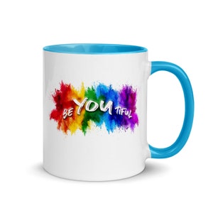 May include: White ceramic coffee mug with a blue rim and handle. The mug features a rainbow-colored explosion of paint with the words "Be You Tiful" in white.
