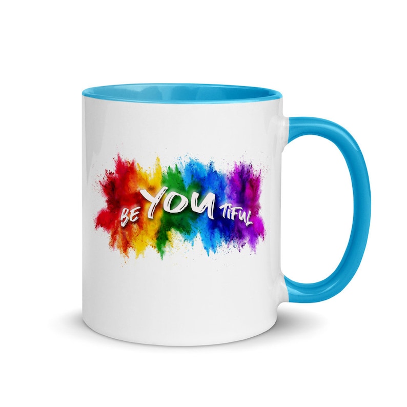May include: White ceramic coffee mug with a blue rim and handle. The mug features a rainbow-colored explosion of paint with the words "Be You Tiful" in white.
