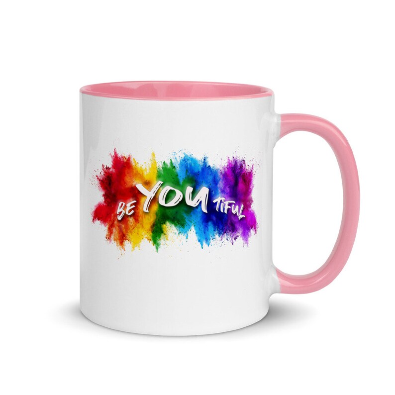 May include: White ceramic mug with a pink rim and handle. The mug features a rainbow-colored paint splatter design with the text "BE YOU TIFUL" in white.