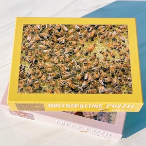 May include: A yellow puzzle box with a close-up image of bees on a honeycomb. The box reads "QUEENSPOTTING PUZZLE" and "1000 PIECES". A pink puzzle box with the text "POPPY PUZZLE" is beneath it.