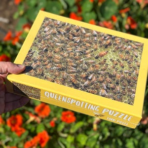 May include: A yellow box puzzle with a close-up image of honeybees. The box reads "QUEENSPOTTING PUZZLE" and "1000 PIECES". The background is blurred with green foliage and orange flowers.