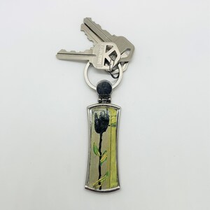 May include: A silver keychain with a black tulip design on a yellow and green background. The keychain has a silver ring and is attached to two keys.