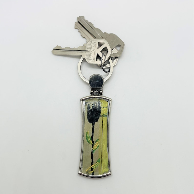 May include: A silver keychain with a black tulip design on a yellow and green background. The keychain has a silver ring and is attached to two keys.