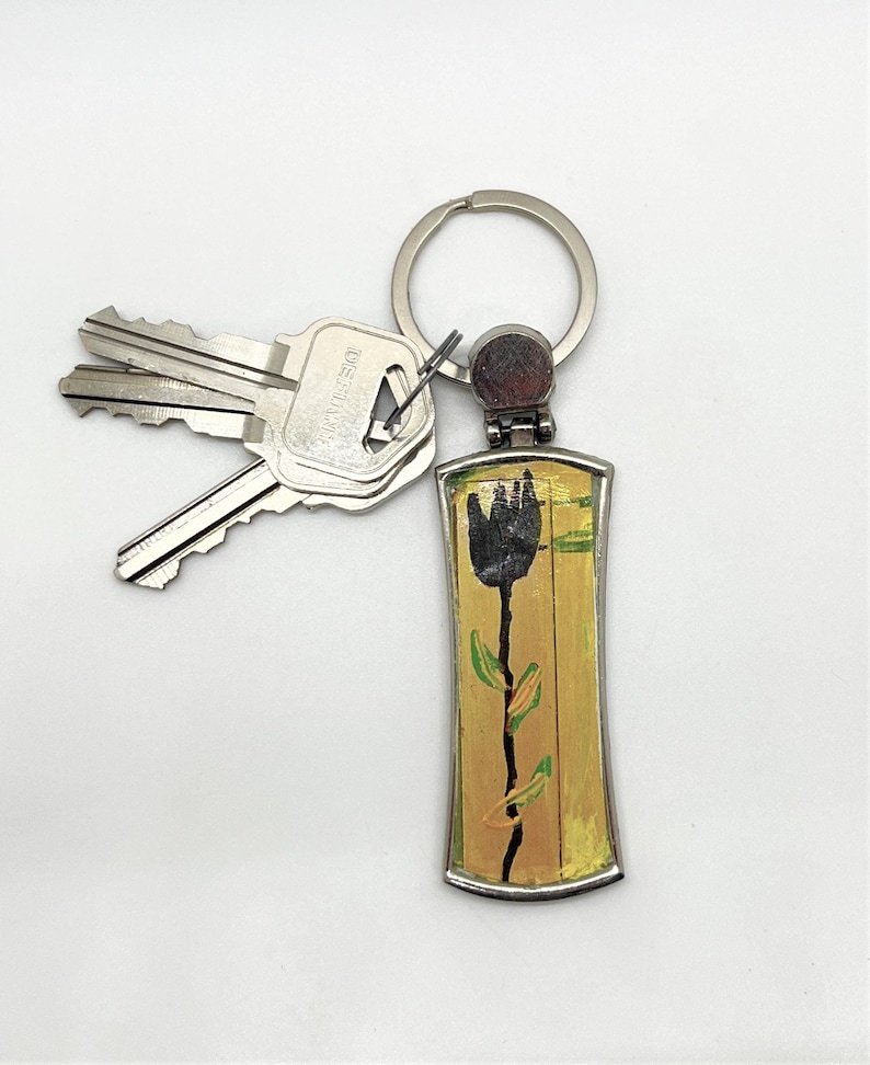 May include: A silver keychain with a black tulip design on a yellow background. The keychain has a silver ring and a silver key attached.