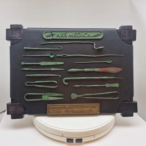 May include: A display board featuring replicas of Roman medical instruments in a verdigris finish mounted on a dark leather-like panel with a wooden frame. The board includes the text ARS MEDICA and Roman Medical Instruments 1st-4th Centuries AD.