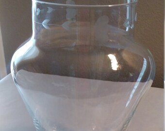 Può includere: Un vaso di vetro trasparente con una base larga e un collo stretto. Il vaso ha un delicato motivo di fiori inciso intorno alla parte superiore.
