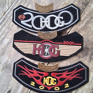 Puede incluir: Tres parches bordados negros con detalles en blanco, rojo y dorado. Cada parche presenta el texto "2000", "2001" y "2002", junto con las letras "HCG". Pequeños emblemas metálicos dorados están encima de cada parche.