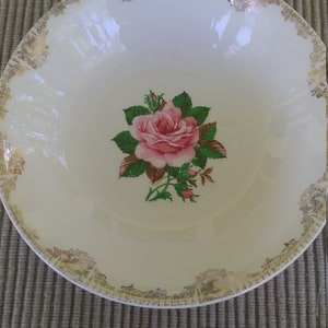 May include: A white ceramic bowl with a gold rim and a pink rose design. The bowl has a scalloped edge and a floral pattern around the rim.