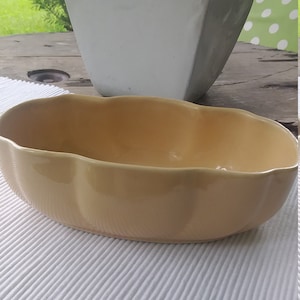Może przedstawiać: Ceramiczna doniczka w jasnożółtym kolorze z falistym brzegiem. Doniczka jest pusta i stoi na białej, teksturowanej powierzchni.