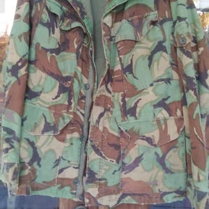 May include: A camouflage pattern jacket in shades of green, brown, and black hangs on a hanger. The jacket has multiple pockets and a zipper closure. The jacket is made of a durable fabric.
