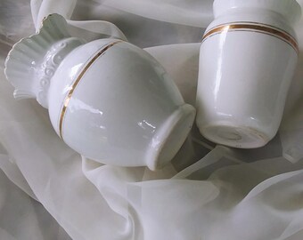 Può includere: Due vasi in ceramica bianca con finiture dorate, uno con la parte superiore arricciata, su uno sfondo di tessuto bianco.