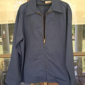 May include: A navy blue zip-up jacket with a collared neckline. The jacket has a gold-colored zipper and a small pocket on the left sleeve. The jacket is hanging on a black hanger.